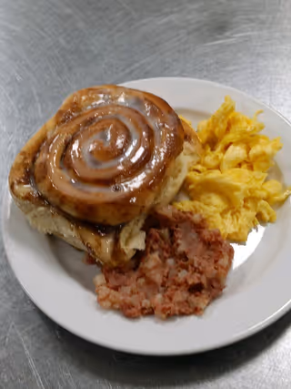 A white plate with a glazed cinnamon roll, scrambled eggs, and a serving of corned beef hash on a metal surface.