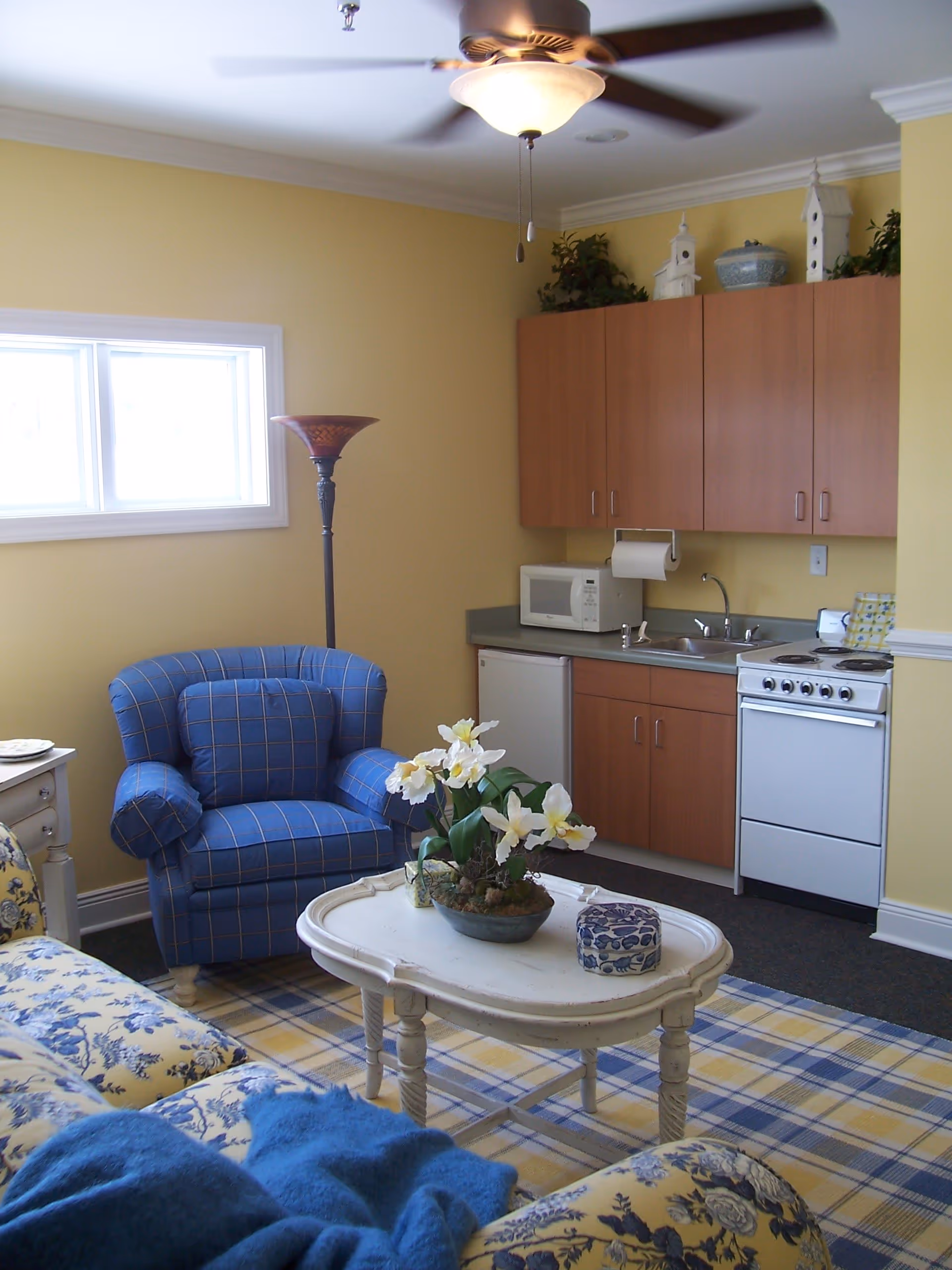 A cozy living area with a blue and yellow floral patterned sofa and a matching blue armchair. A white coffee table with a flower arrangement sits on a yellow and blue plaid rug. The room has yellow walls, a ceiling fan with a light, and a small kitchenette with wooden cabinets, a microwave, a mini fridge, a sink, and a stove. A tall floor lamp stands near the armchair.