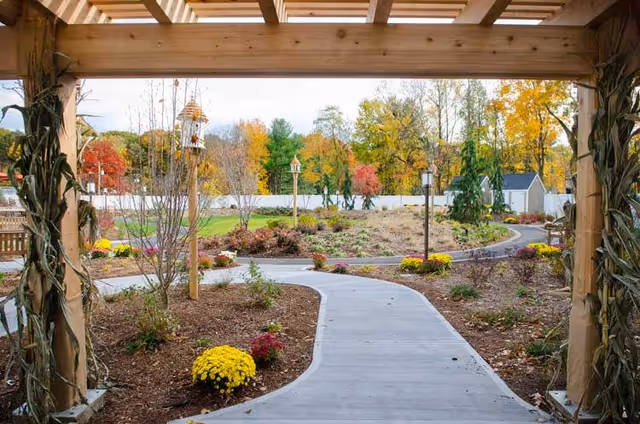 View of a winding concrete pathway through a landscaped garden with colorful autumn foliage, flower beds, birdhouses, and a small shed in the background, framed by a wooden pergola decorated with dried corn stalks.