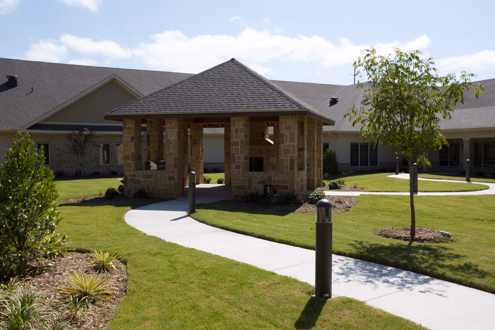Outdoor view of a landscaped garden area with a stone pavilion structure featuring a shingled roof, surrounded by a curved concrete walkway, grass, small trees, and shrubs. Residential-style buildings are visible in the background under a partly cloudy sky.