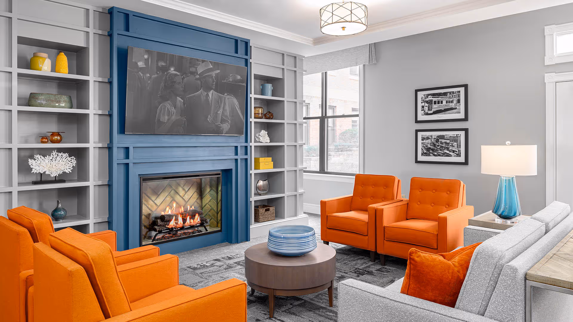 A cozy living room with bright orange armchairs and a light gray sofa with orange pillows arranged around a round wooden coffee table. The room features a blue fireplace with a fire burning and a black-and-white movie playing on a TV mounted above it. Built-in shelves flank the fireplace, decorated with various vases and decorative items. A large window lets in natural light, and two black-and-white framed photos hang on the gray wall next to a table lamp with a blue base.