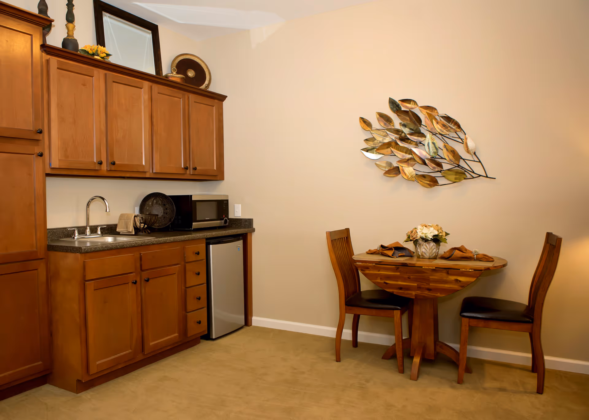 Small kitchenette with wooden cabinets, sink, microwave, mini fridge and a round wooden table with two chairs beneath a metal leaf wall decoration.