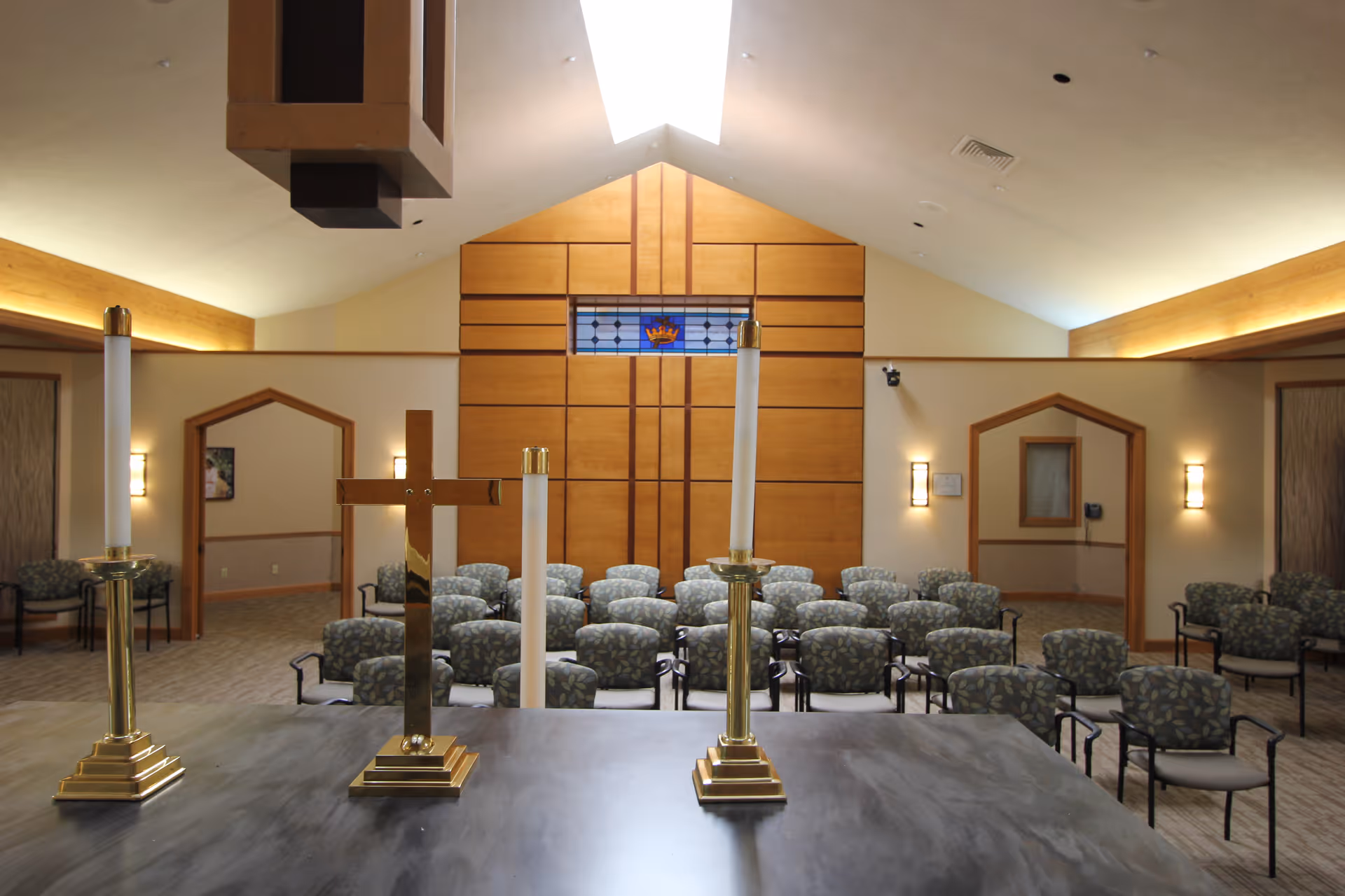 Chapel interior with an altar holding a brass cross and candlesticks, and rows of patterned chairs facing a wood-paneled wall with a stained-glass window.