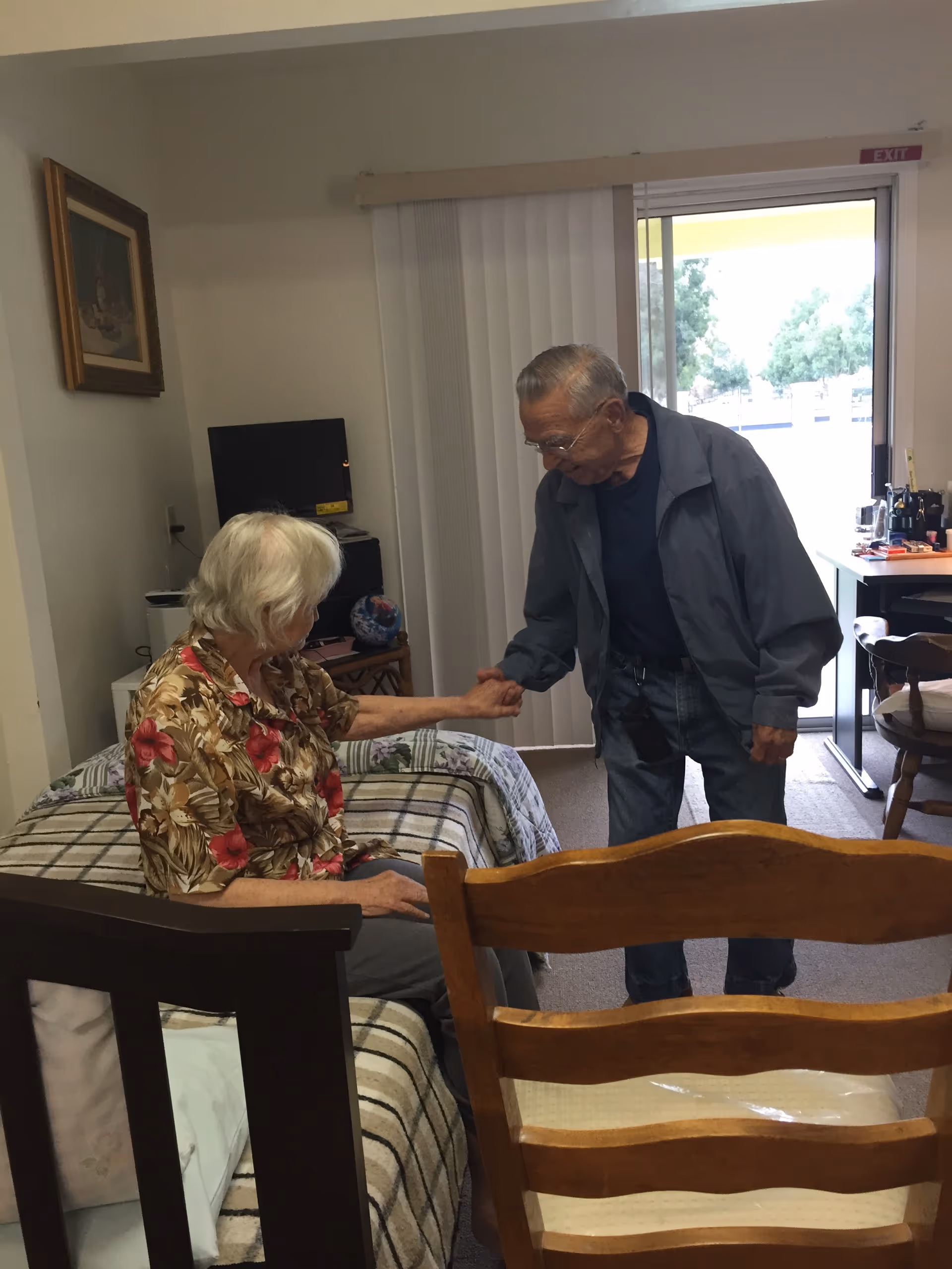 An elderly woman sitting on a bed inside a room, holding hands with an elderly man standing beside her. The room has a bed with a striped blanket, a wooden chair in the foreground, a desk with items near a sliding glass door with vertical blinds, and a framed picture on the wall.