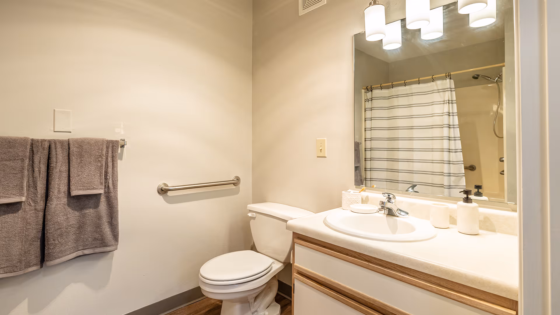 A clean bathroom with a toilet, a sink with a countertop, a large mirror above the sink, a striped shower curtain, two gray towels hanging on a towel rack, and a grab bar on the wall next to the toilet.