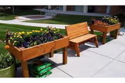 Outdoor patio area with two wooden raised garden beds filled with blooming flowers and a wooden bench placed between them on a concrete sidewalk. Green grass and a building entrance are visible in the background.
