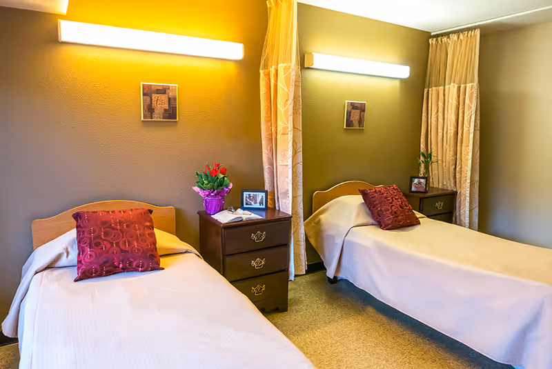 A room with two single beds separated by a wooden nightstand. Each bed has a beige bedspread and a red decorative pillow. The nightstand holds a small potted plant with red flowers and a framed photo. The walls are painted in a warm yellow and beige tone, with two wall-mounted lights above each bed and curtains hanging beside each bed.