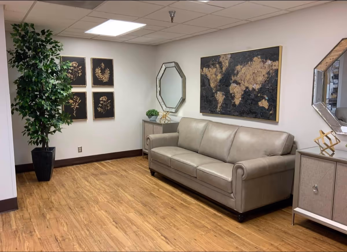 A cozy interior room with a light gray leather sofa against a white wall. Above the sofa is a large framed world map artwork. On either side of the sofa are matching light-colored cabinets with decorative items and octagonal mirrors above them. To the left, there is a tall green potted plant and four framed botanical prints on the wall. The floor is wood with a warm tone, and the ceiling has recessed lighting.