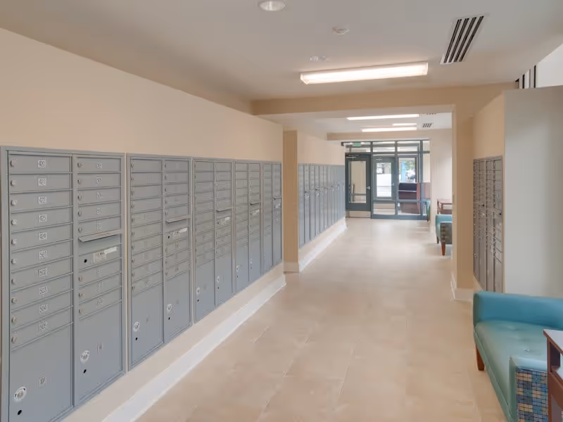 Interior hallway of Linden Park Apartments in Bolton Hill featuring rows of gray mailboxes mounted on the walls on both sides, beige tiled floor, beige walls, ceiling lights, and a seating area with teal armchairs near the glass entrance door at the far end.