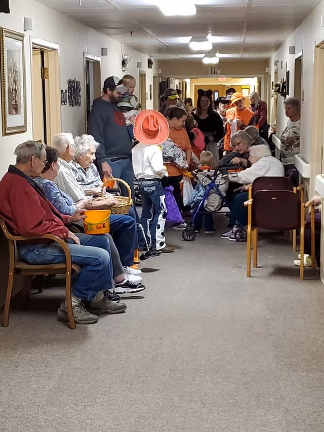 A hallway in a senior living facility with elderly residents seated along one side and children dressed in Halloween costumes walking down the hall. Some adults are standing among the children, and the atmosphere appears festive and social.