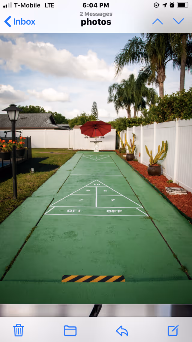 Outdoor shuffleboard court with green painted surface and white markings, surrounded by grass and a white fence. There are potted plants with cacti along the right side and a red umbrella at the far end near a small table. Palm trees and a partly cloudy sky are visible in the background.