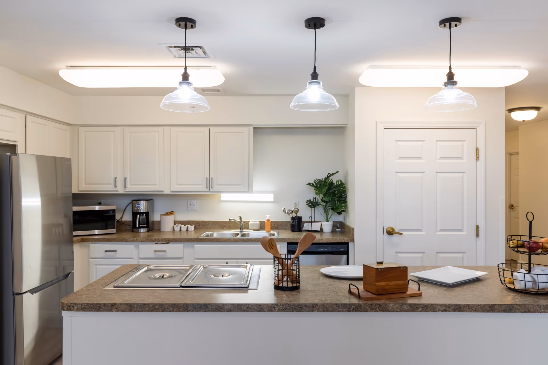 A modern kitchen with white cabinets, a stainless steel refrigerator, a microwave, a coffee maker, and a double sink. The countertop has a built-in stove with two covered burners, wooden cooking utensils in a holder, plates, a wooden box, and a fruit basket. Three pendant lights hang from the ceiling, illuminating the space.