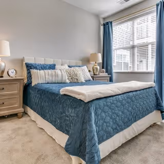 A bright bedroom with a bed dressed in blue bedding, matching curtains, nightstands and lamps by a large window.