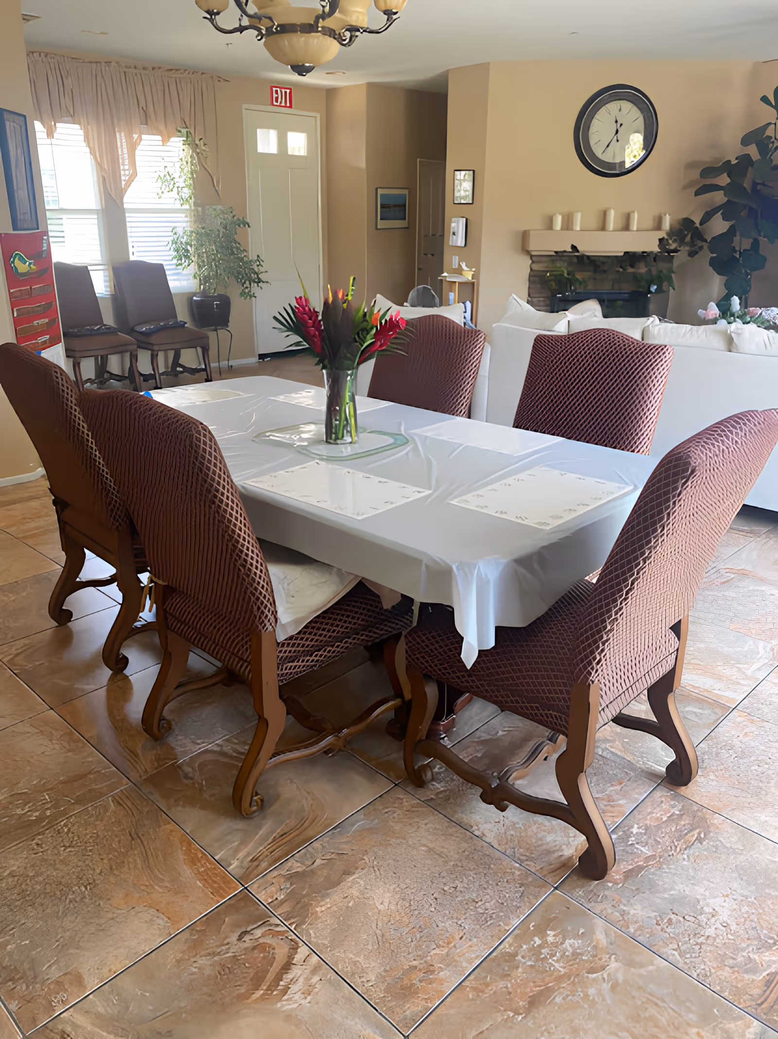 A dining area with a rectangular table covered with a white tablecloth and six chairs with brown patterned covers. A vase with red and green flowers is placed in the center of the table. In the background, there is a white couch, a fireplace with candles on the mantel, a large clock on the wall, and some plants near the windows.