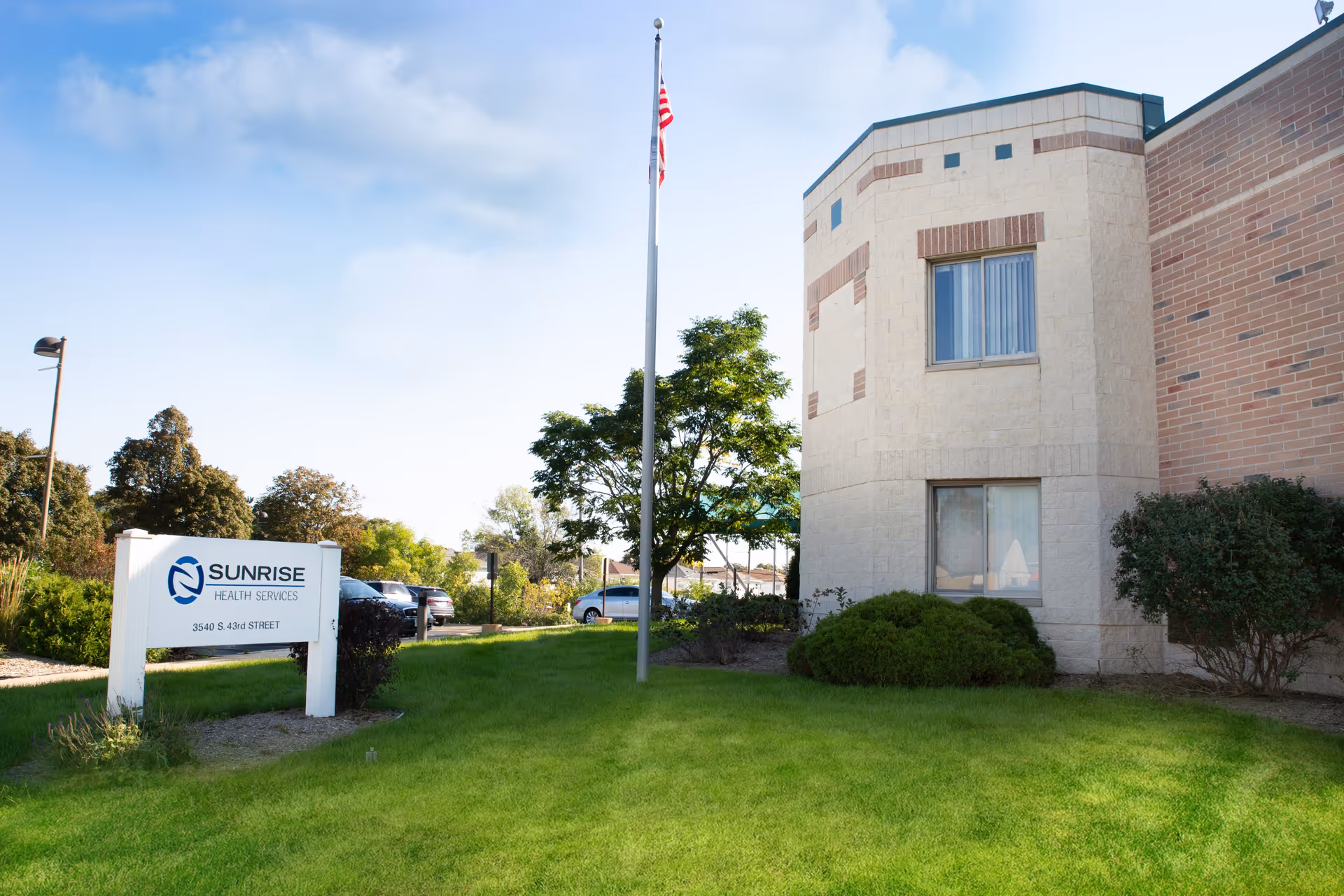 Exterior view of Sunrise Health Services building with a green lawn, bushes, a flagpole with an American flag, and a sign displaying the facility's name and address.