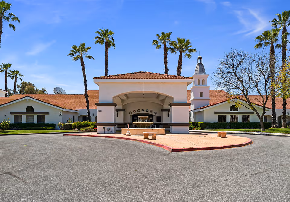 Front exterior view of Westmont of Riverside facility with a circular driveway, a covered entrance with a fountain, palm trees, and a clear blue sky.
