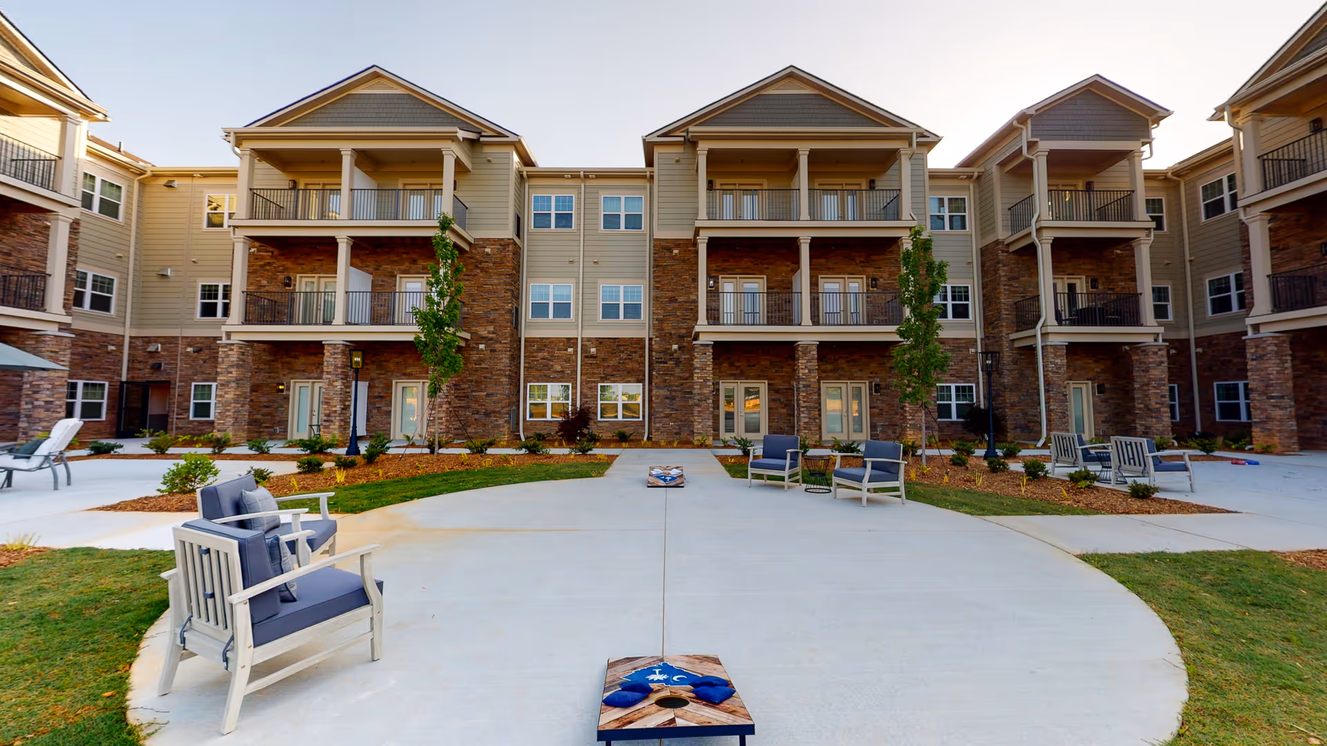 Outdoor courtyard area of a senior living facility with a concrete patio featuring a cornhole game setup, surrounded by cushioned wooden chairs and small tables. The background shows a three-story building with balconies, stone and siding exterior, and landscaped greenery.