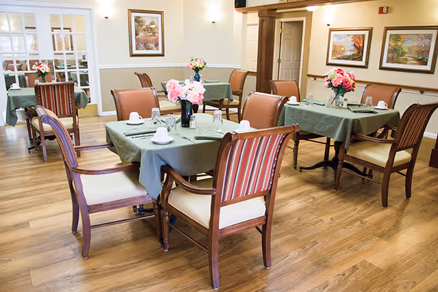 A dining room with several tables covered in green tablecloths, each set with white cups, glasses, and silverware. Wooden chairs with cushioned seats surround the tables. The room has wooden flooring, framed artwork on the walls, and floral centerpieces on each table.