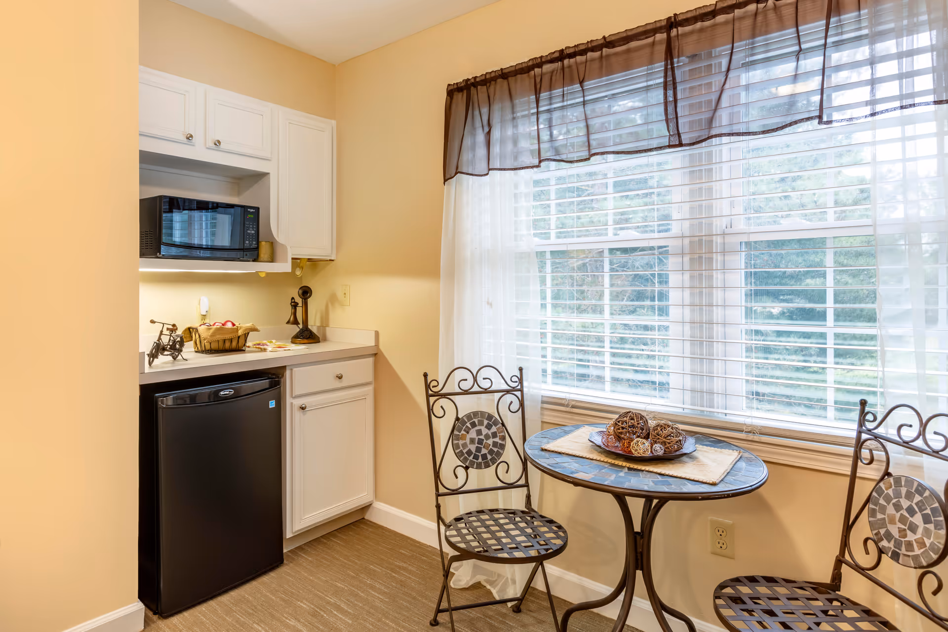 A small kitchen nook with a black mini refrigerator, a microwave on a white cabinet, and a countertop with decorative items. Next to the kitchen area is a round table with a mosaic top and two wrought iron chairs with decorative backs, placed in front of a large window with white blinds and sheer curtains.
