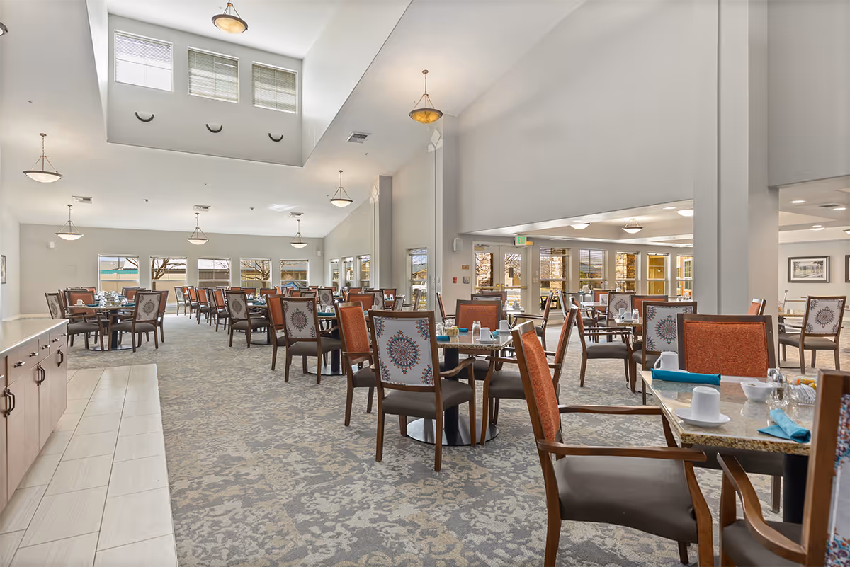 Spacious dining room in a senior living facility with multiple tables and chairs arranged neatly. The room has high ceilings with hanging light fixtures, large windows allowing natural light, and a patterned carpet floor. Tables are set with cups, napkins, and condiments.