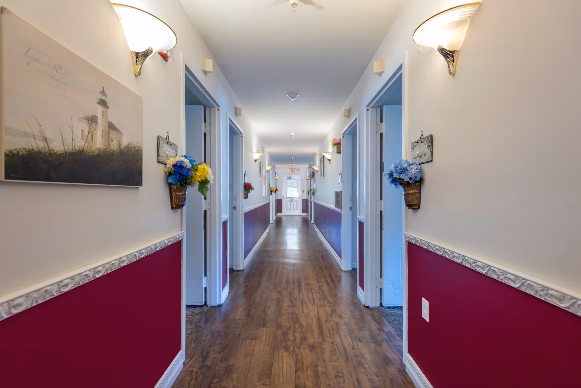 A long, well-lit hallway in a senior living facility with wooden flooring and walls painted white on the upper half and dark red on the lower half. Several open doorways line both sides of the hallway. Wall-mounted light fixtures and decorative flower arrangements in small wall baskets are evenly spaced along the corridor. A painting of a lighthouse hangs on the left wall near the foreground.