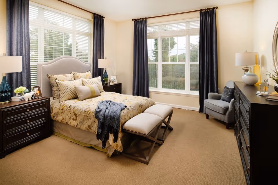 A cozy bedroom with a large bed adorned with multiple pillows and a floral bedspread. The room features two dark wooden nightstands with lamps, a cushioned bench at the foot of the bed, a comfortable armchair, a dark wooden dresser with decorative items, and large windows with dark curtains allowing natural light to fill the space.