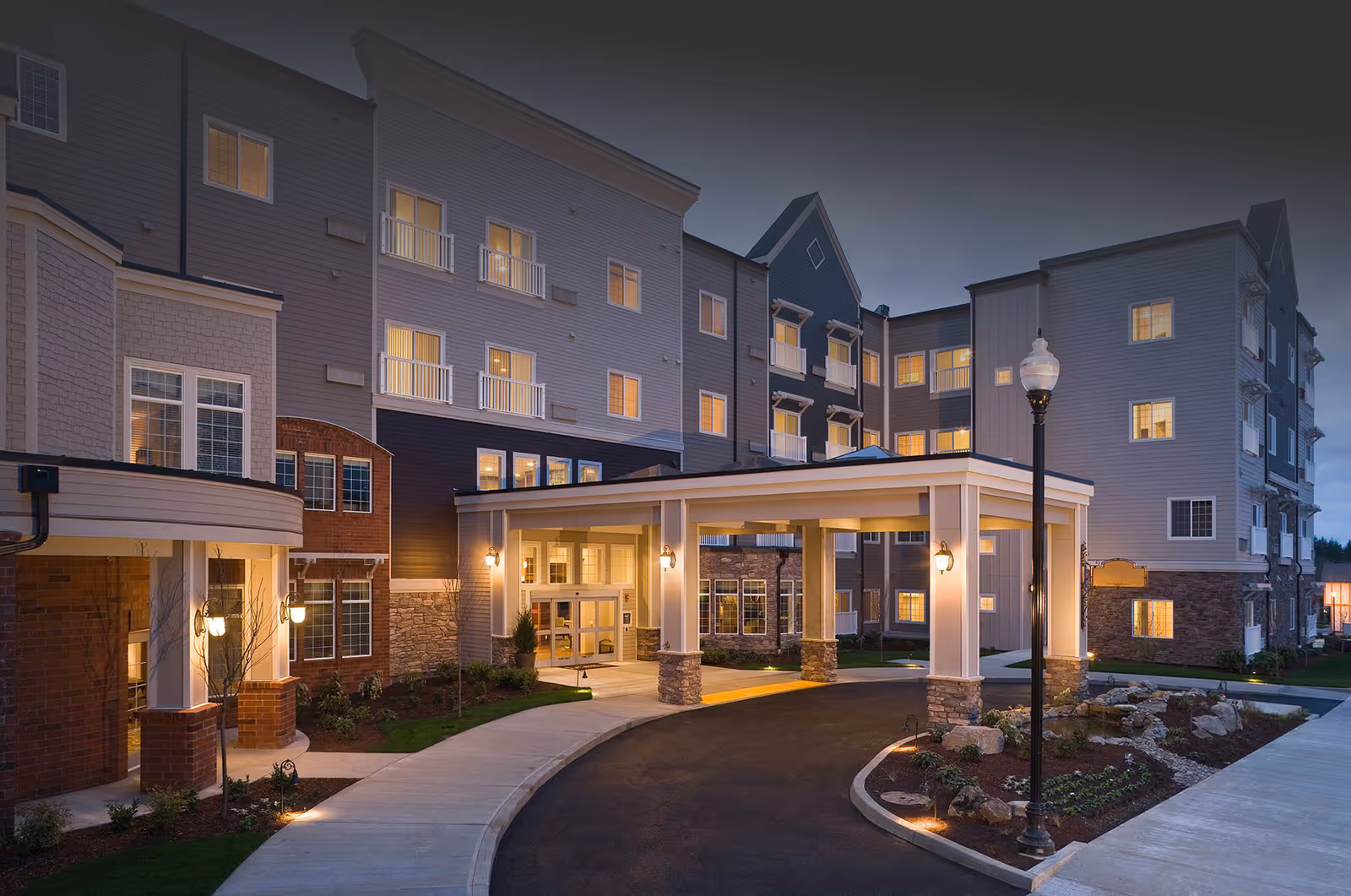 Exterior view of a senior living community building at dusk with lights on inside. The building has multiple floors with windows and balconies. There is a covered entrance with stone pillars and a curved driveway leading up to it. Landscaping with small plants and rocks is visible along the driveway.