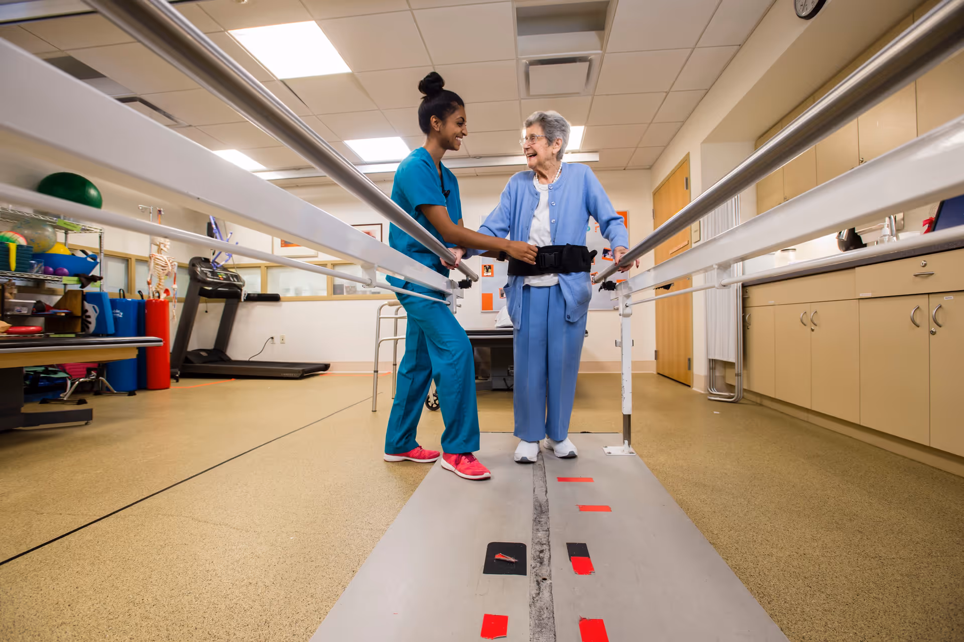 A healthcare worker in blue scrubs assists an elderly woman using parallel bars for walking practice in a rehabilitation or physical therapy room. The elderly woman is smiling and wearing a blue cardigan and pants with a gait belt. The room has exercise equipment and cabinets along the walls.
