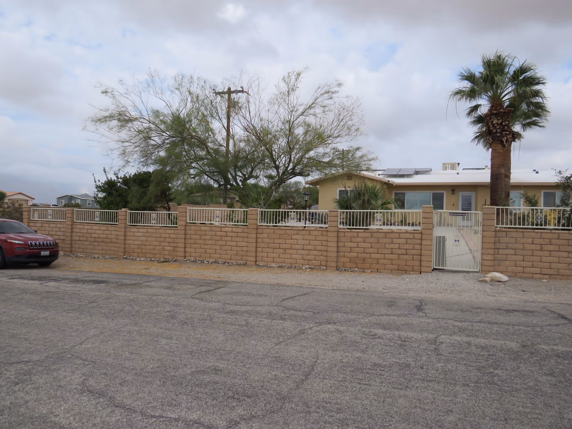 Exterior view of a single-story building behind a beige brick wall with a white metal gate. There is a tall palm tree and other greenery visible inside the fenced area. A red vehicle is parked on the street in front of the wall under a cloudy sky.