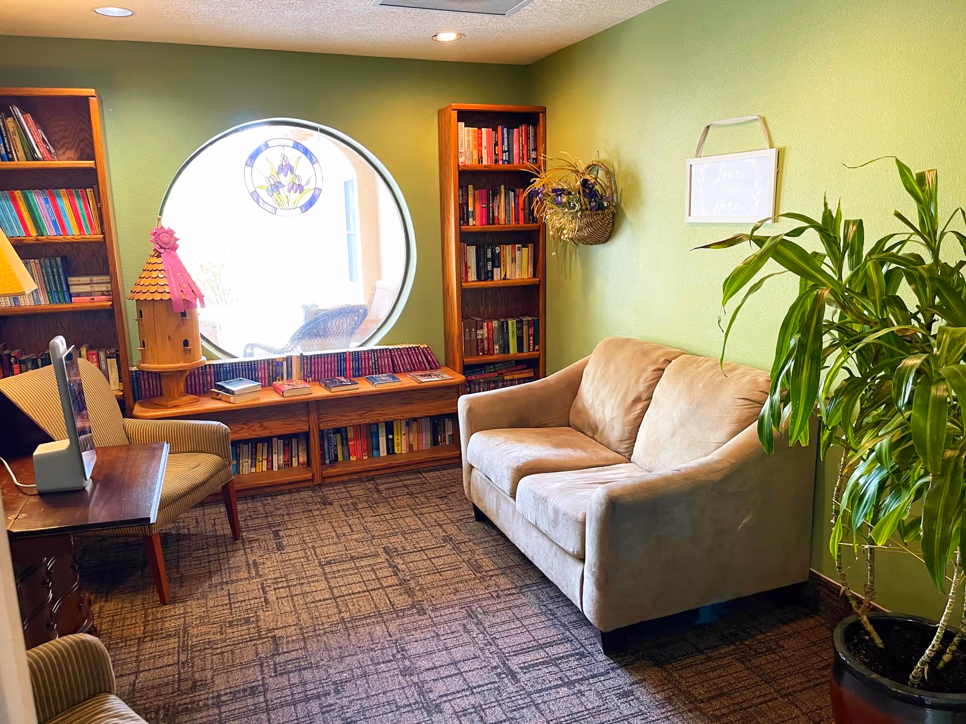 A cozy reading nook in a senior living facility with a beige loveseat, two armchairs, wooden bookshelves filled with books, a round window with a stained glass decoration, a birdhouse, and a large potted plant against a green wall.