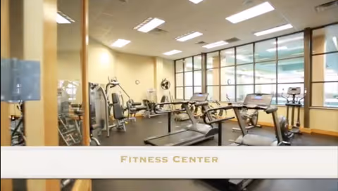 Interior view of a fitness center with exercise equipment including treadmills, stationary bikes, and weight machines. The room has large windows allowing natural light to enter and a light-colored ceiling with recessed lighting.