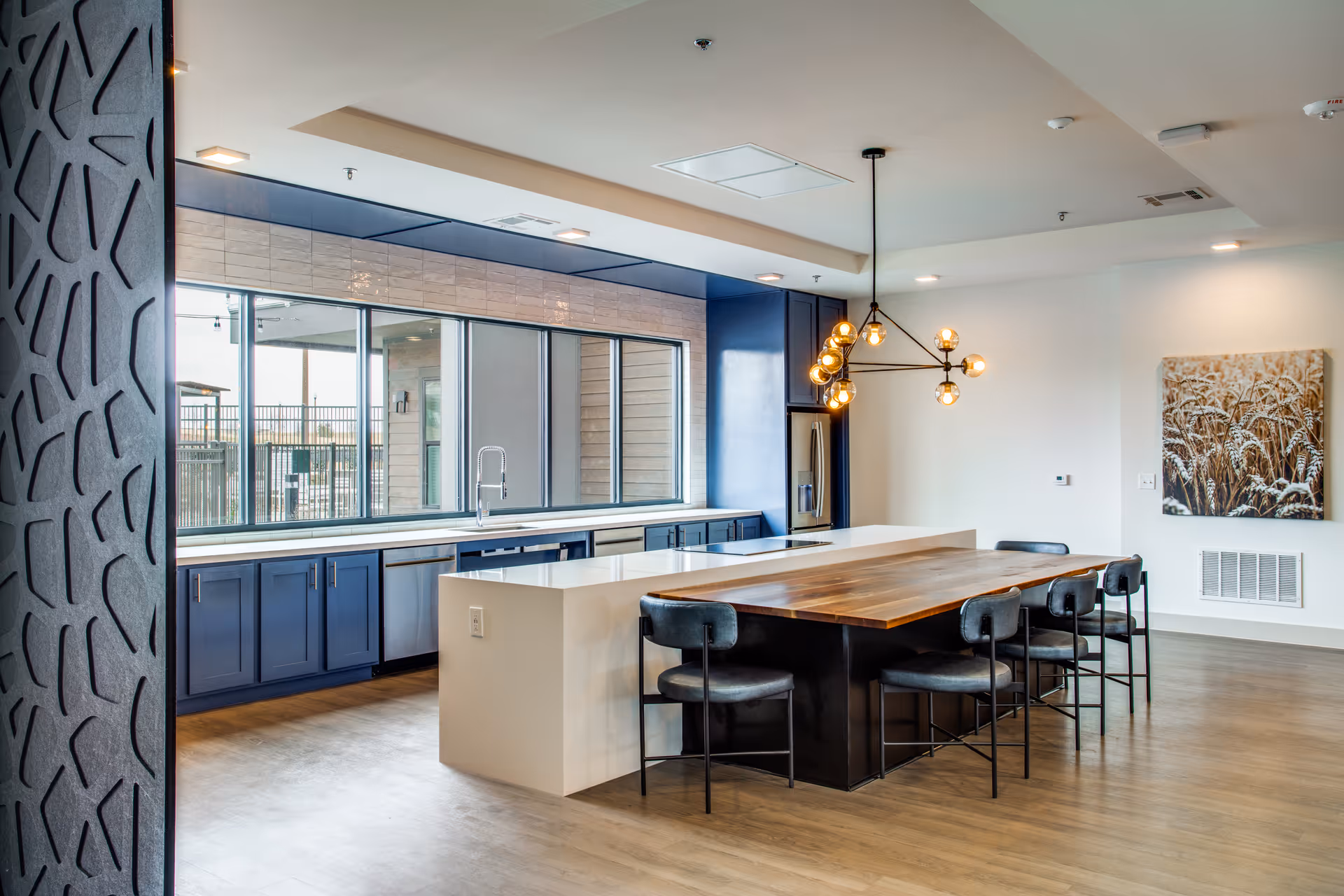 Modern kitchen area with a large wooden dining table surrounded by black chairs, a white kitchen island, blue cabinetry, stainless steel refrigerator, large windows letting in natural light, and a contemporary chandelier hanging above the table.
