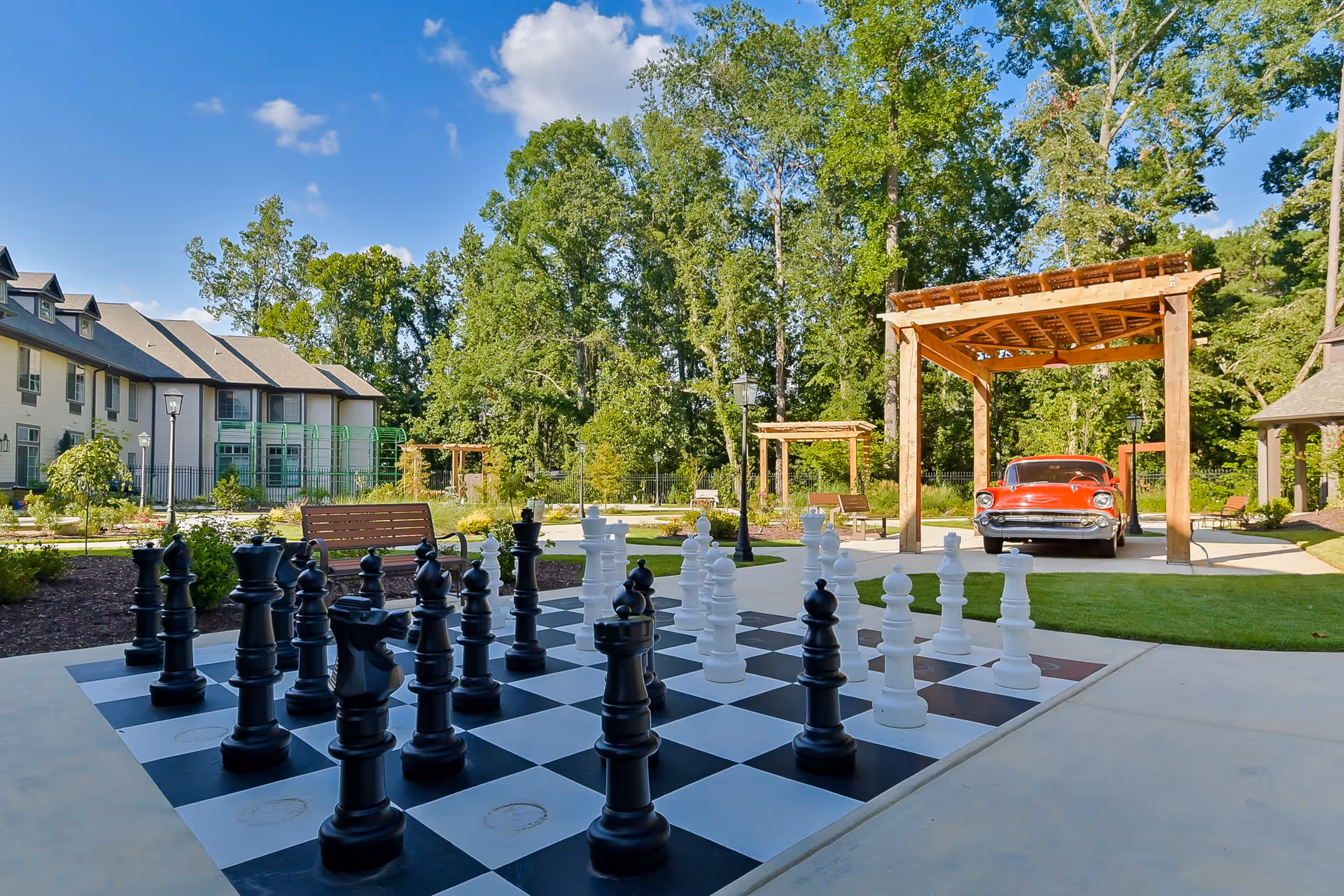 Outdoor courtyard with a life-size chessboard, benches, wooden pergolas, a red vintage car, and apartment buildings surrounded by trees.