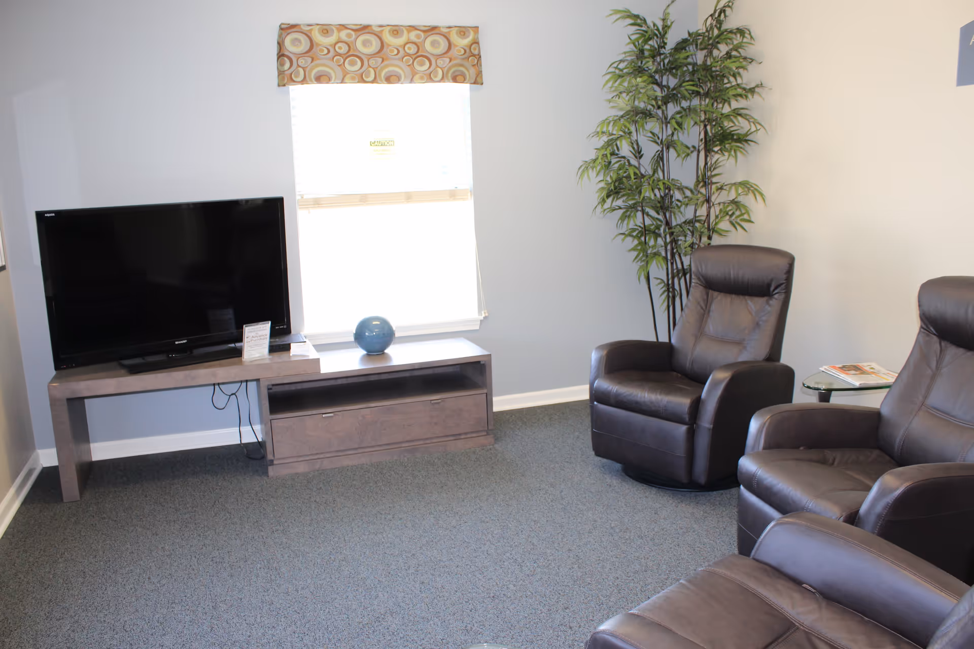 A cozy living room area with three dark brown leather recliner chairs arranged around a flat-screen TV on a wooden TV stand. There is a window with a patterned valance behind the TV, a blue decorative vase on the TV stand, a tall green plant in the corner, and a small glass side table with magazines.