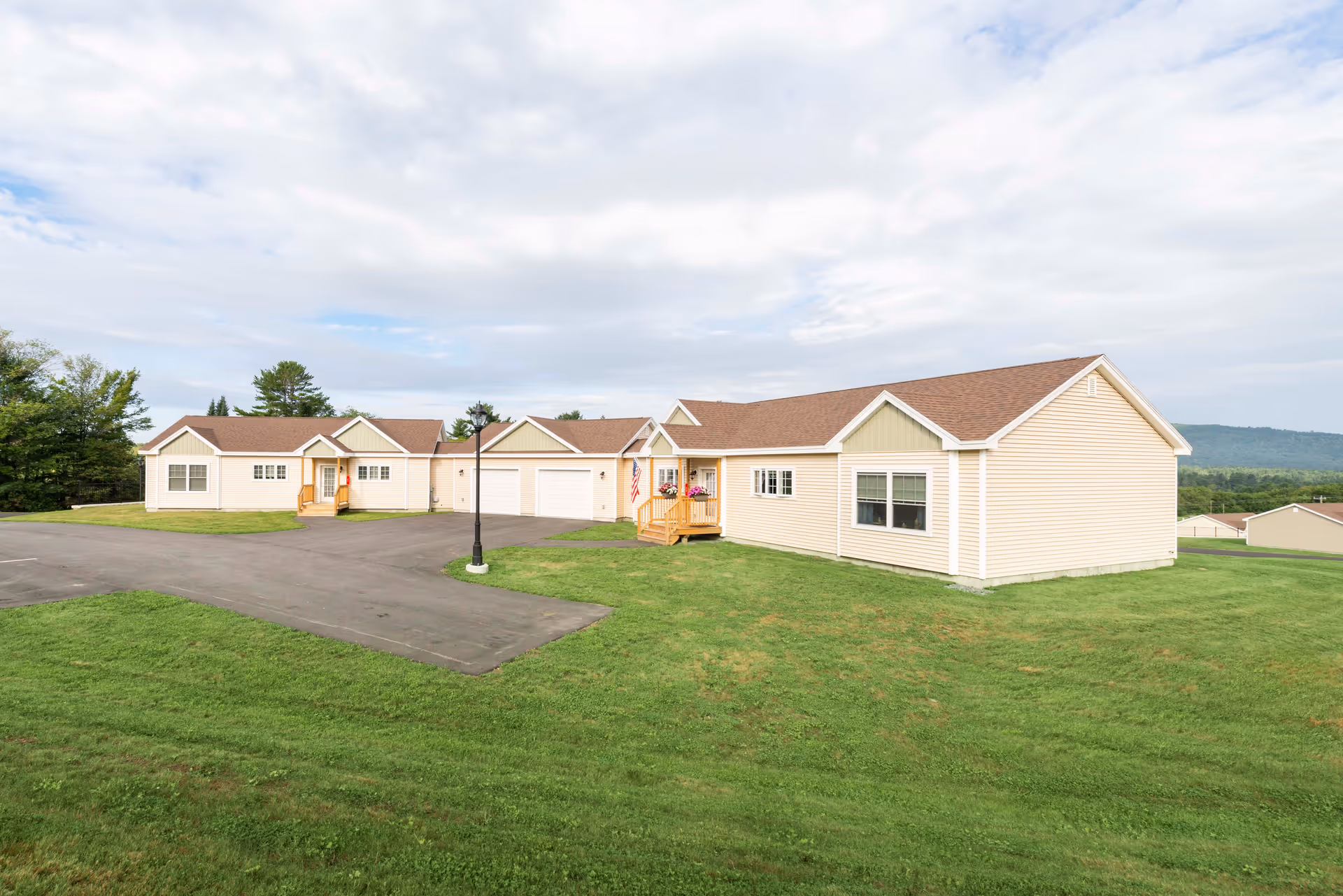 Exterior view of a single-story beige building with brown roof shingles, multiple windows, and small porches with steps leading to the entrances. The building is surrounded by green grass and a paved driveway, with trees and hills visible in the background under a partly cloudy sky.