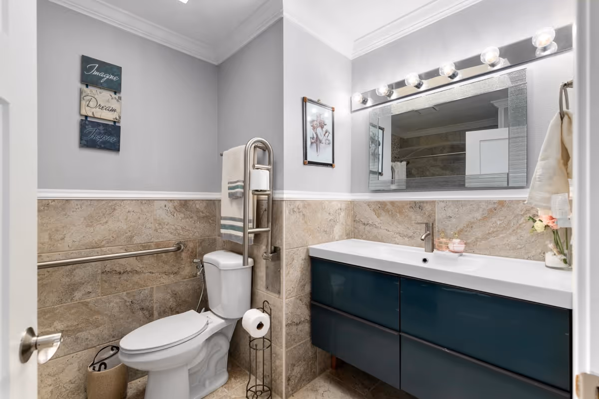 A clean and modern bathroom with beige tiled walls and floor. There is a white toilet with a metal grab bar beside it and a towel rack above. A dark blue vanity with a white countertop and a single faucet is on the right side, with a large mirror above it framed by lights. The bathroom also features wall art and a small vase with flowers on the countertop.
