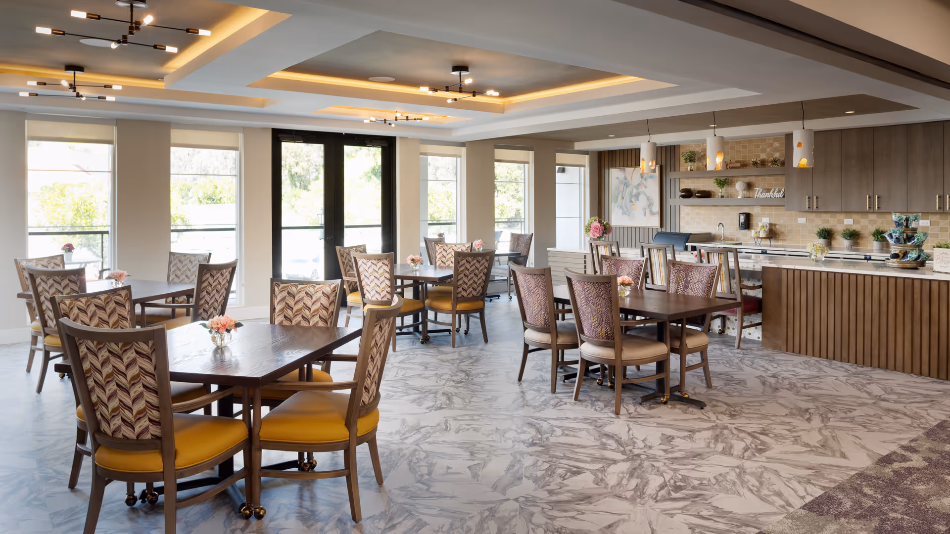 Bright, spacious dining room with several wooden tables and upholstered chairs, large windows, and a service counter/kitchen area.