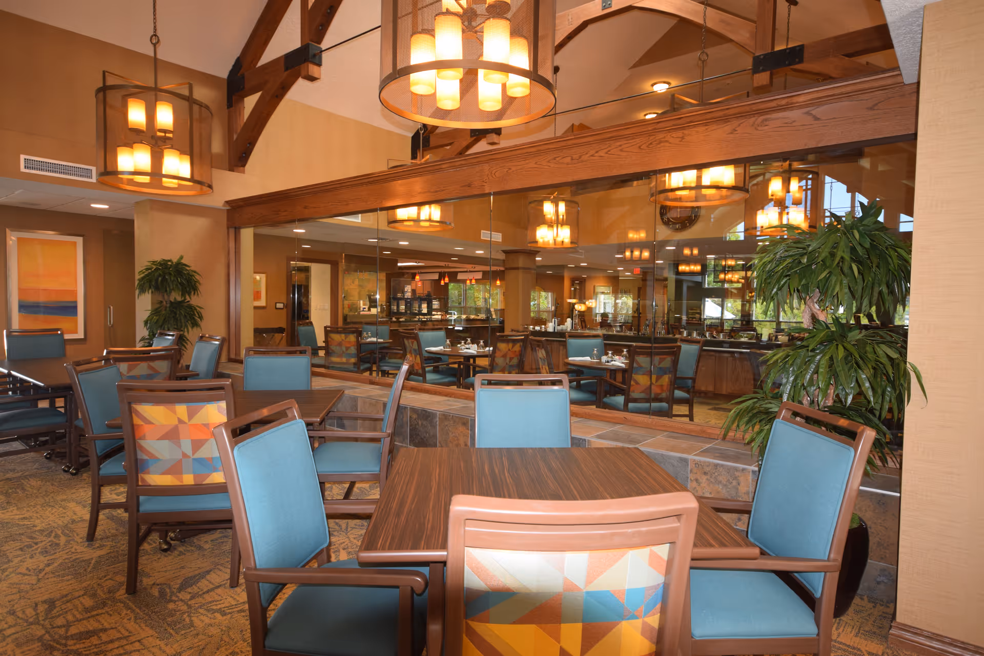 A dining area in a retirement community with wooden tables and chairs featuring blue and patterned upholstery. The room has warm lighting with hanging pendant lights and a large mirror reflecting the space. There are plants and framed artwork on the walls, creating a cozy and inviting atmosphere.