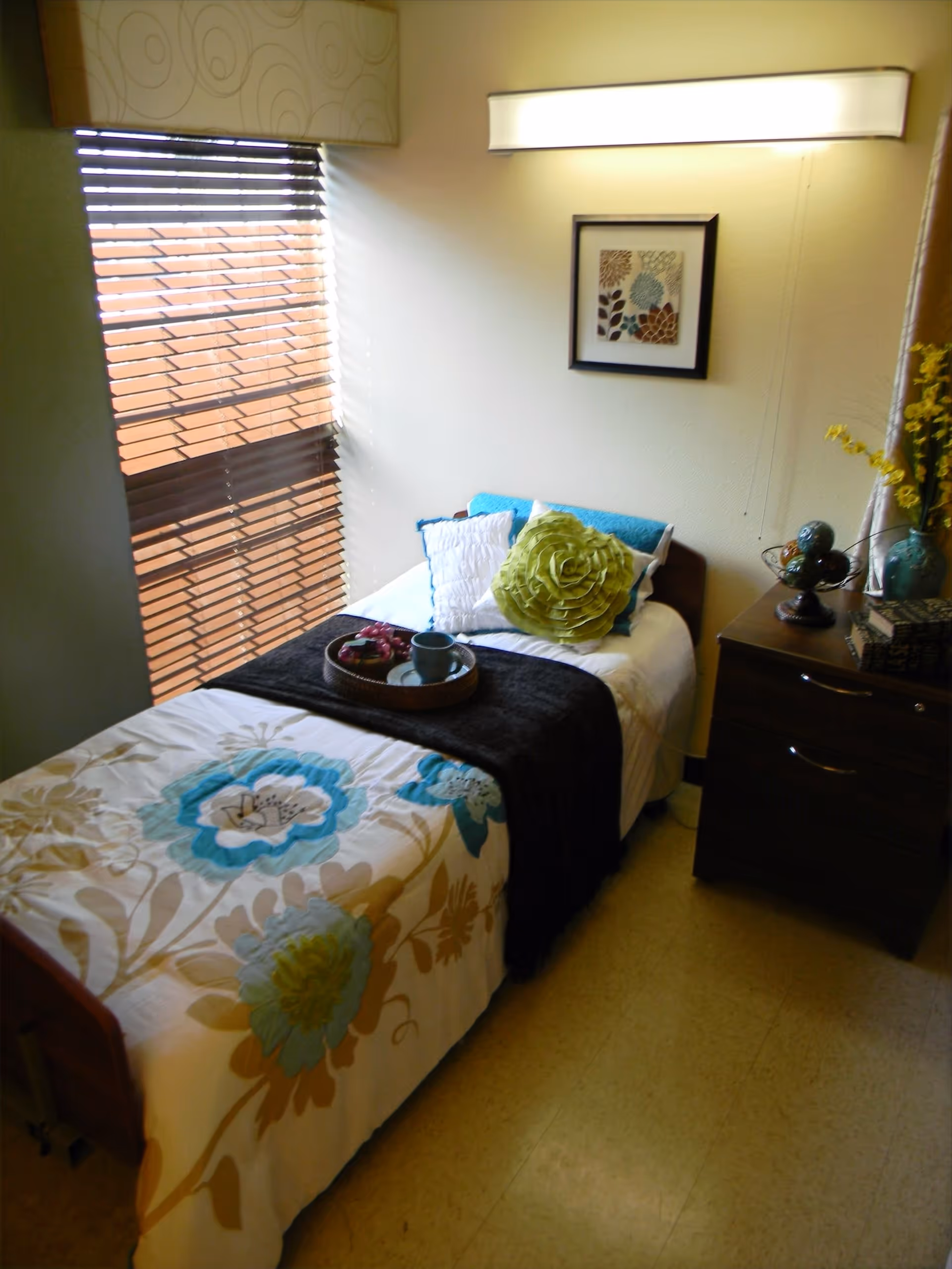 A small, cozy bedroom with a single bed covered in a floral-patterned bedspread and decorative pillows, including a large green flower-shaped pillow. A tray with a cup and some fruit is placed on the bed. Next to the bed is a dark wooden nightstand with decorative items and a vase with yellow flowers. A framed floral artwork hangs on the wall above the bed, and a window with wooden blinds lets in natural light.