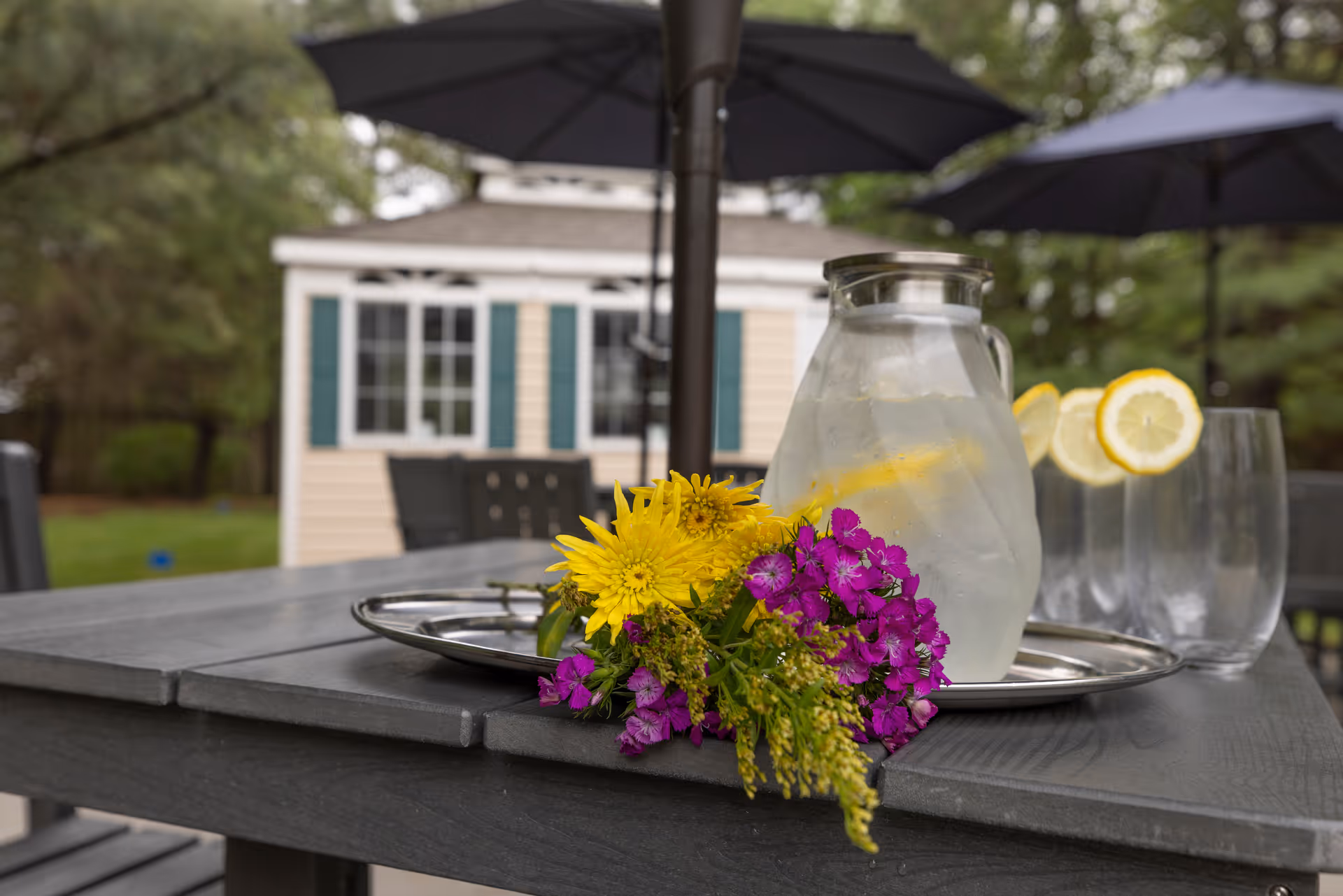 Outdoor table with a silver tray holding a glass pitcher of ice water with lemon slices and two glasses garnished with lemon slices, accompanied by a small bouquet of yellow and purple flowers. In the background, there is a small building with green shutters and large umbrellas providing shade.