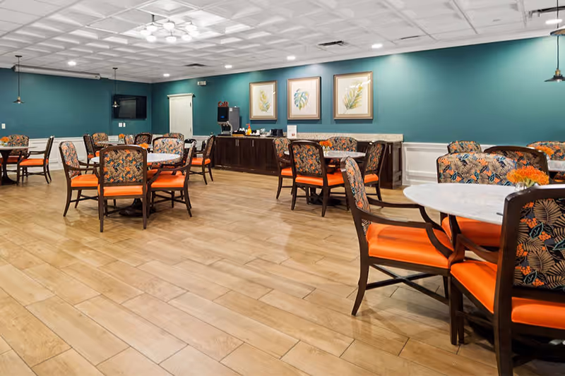 A dining room with several round tables and chairs featuring orange cushions and patterned backs. The room has wooden flooring, teal walls, and three framed botanical prints on one wall above a dark wooden sideboard with a coffee machine and cups. The ceiling has recessed lighting and a white coffered design.