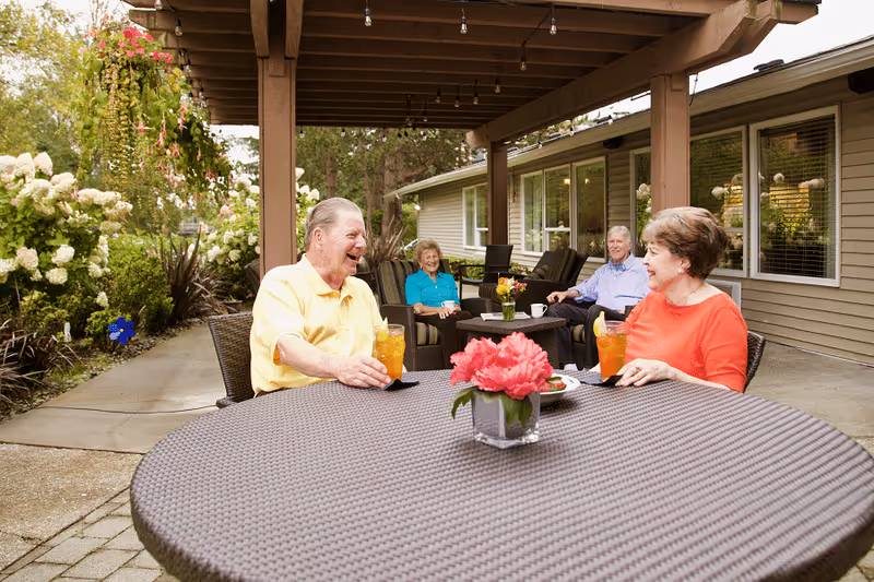 Four elderly people sitting outdoors under a wooden pergola at a senior living facility. Two are seated at a round table with drinks and a flower centerpiece, while the other two sit on chairs in the background near the building. The area is surrounded by greenery and flowers.