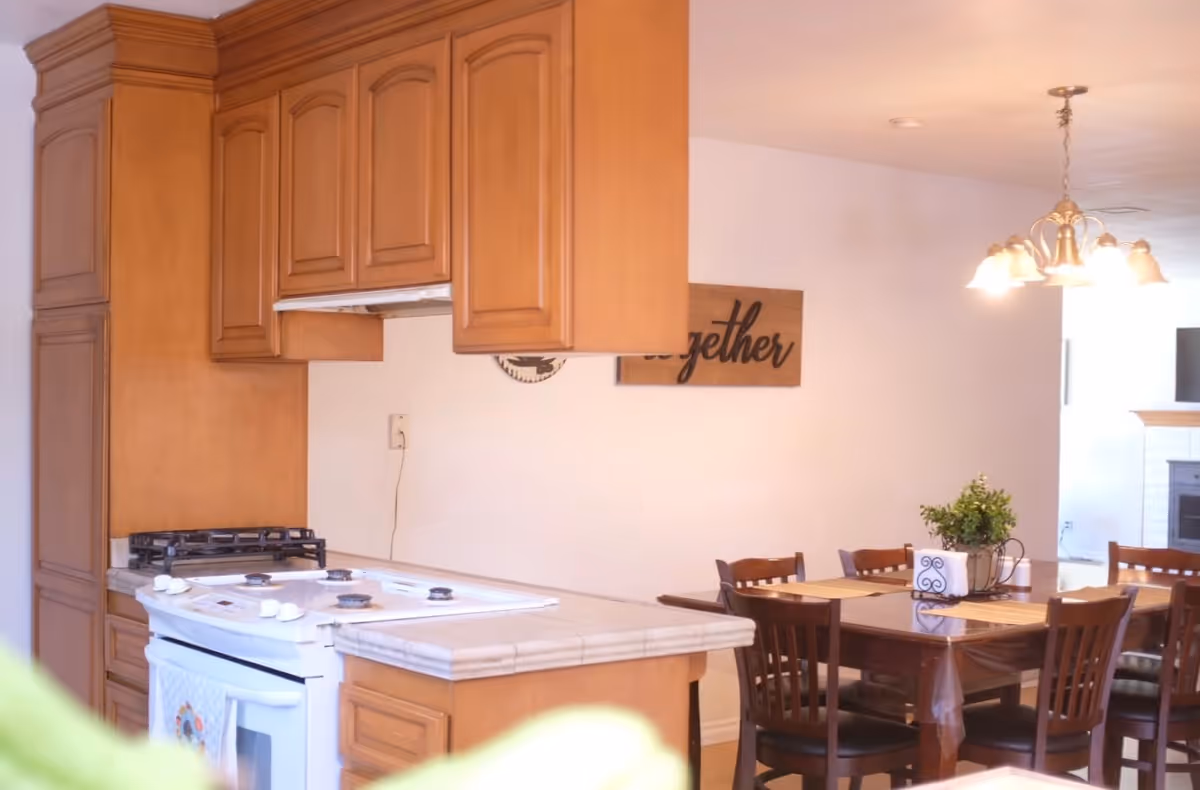 Open kitchen with wooden cabinets and a stove opening onto a dining area with a table, chairs, and a hanging light fixture.