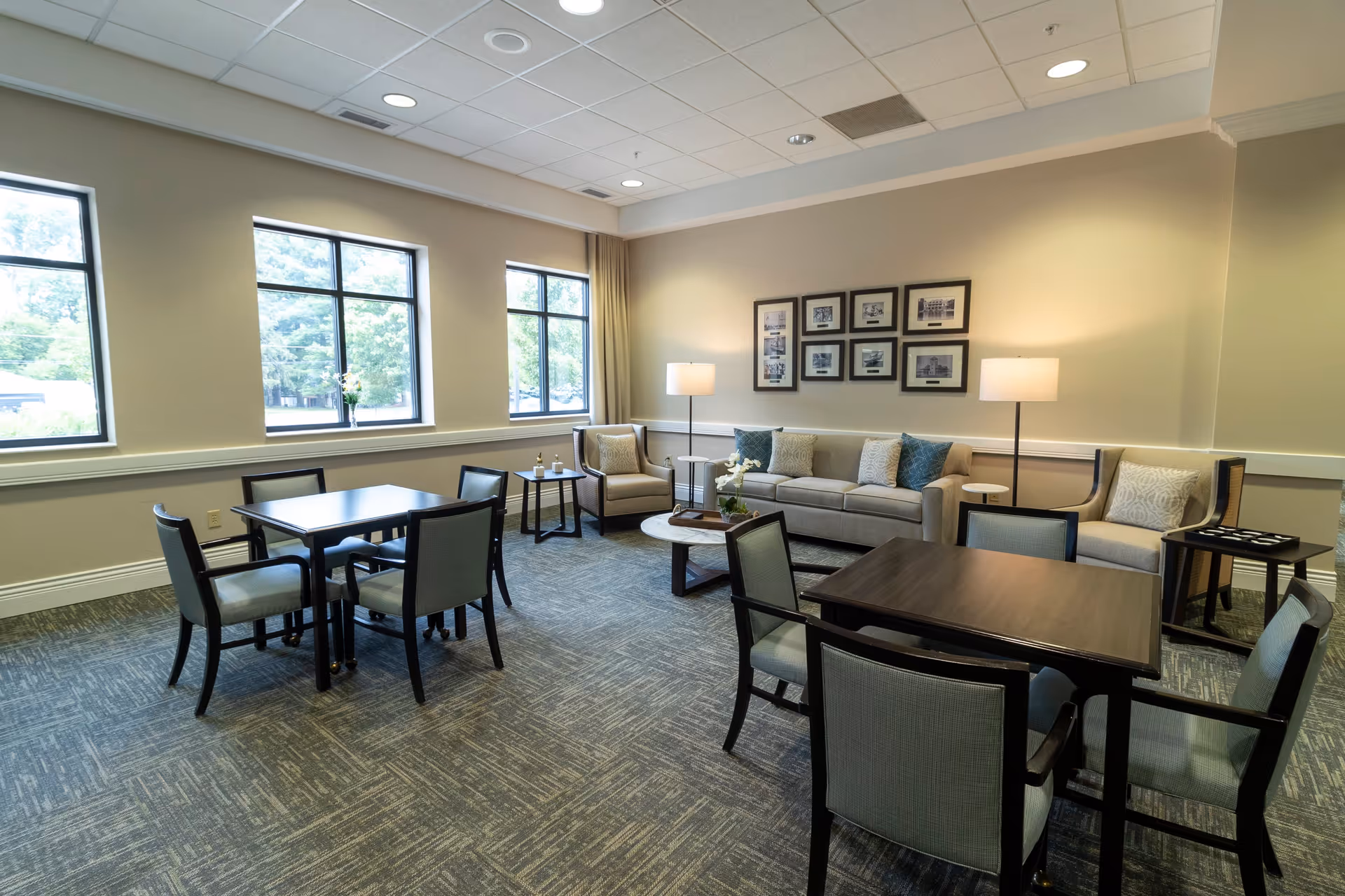 Bright communal lounge with sofas, armchairs, framed photos, and several card tables and chairs by large windows.