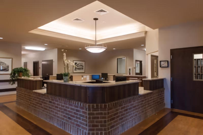 Reception area inside a healthcare and rehabilitation center with a brick and wood front desk, computer monitors, a hanging light fixture, framed pictures on the walls, and a potted plant on the desk.
