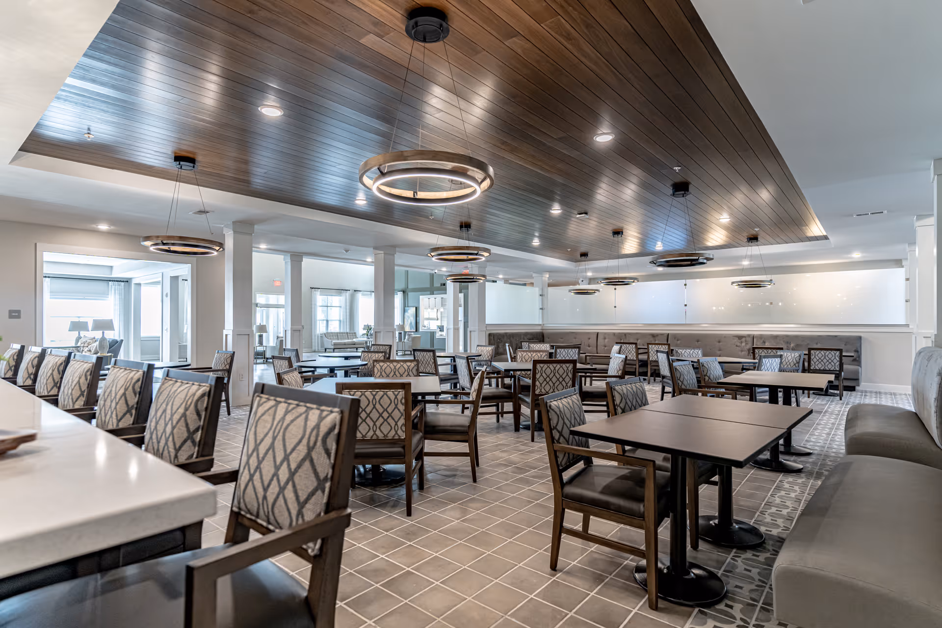 A spacious dining room with multiple tables and chairs arranged neatly. The ceiling features a wood panel design with modern circular pendant lights. The room has a tiled floor and a combination of cushioned bench seating along the walls and individual chairs around the tables. Large windows allow natural light to enter the space.