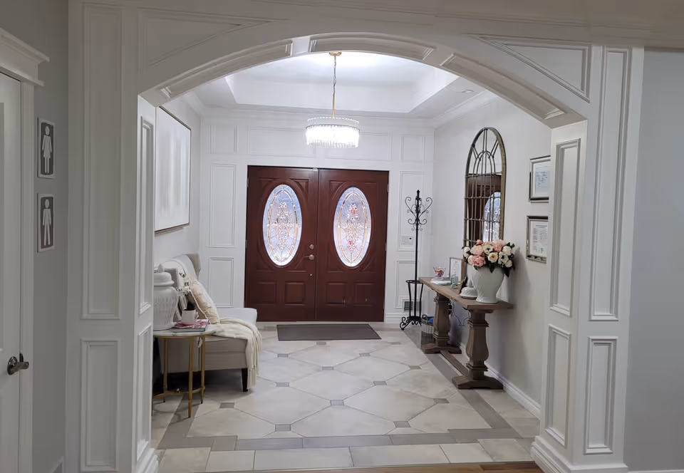 Bright, elegant entry foyer with double wooden front doors, tiled floor, a bench to the left and a console table with flowers to the right.