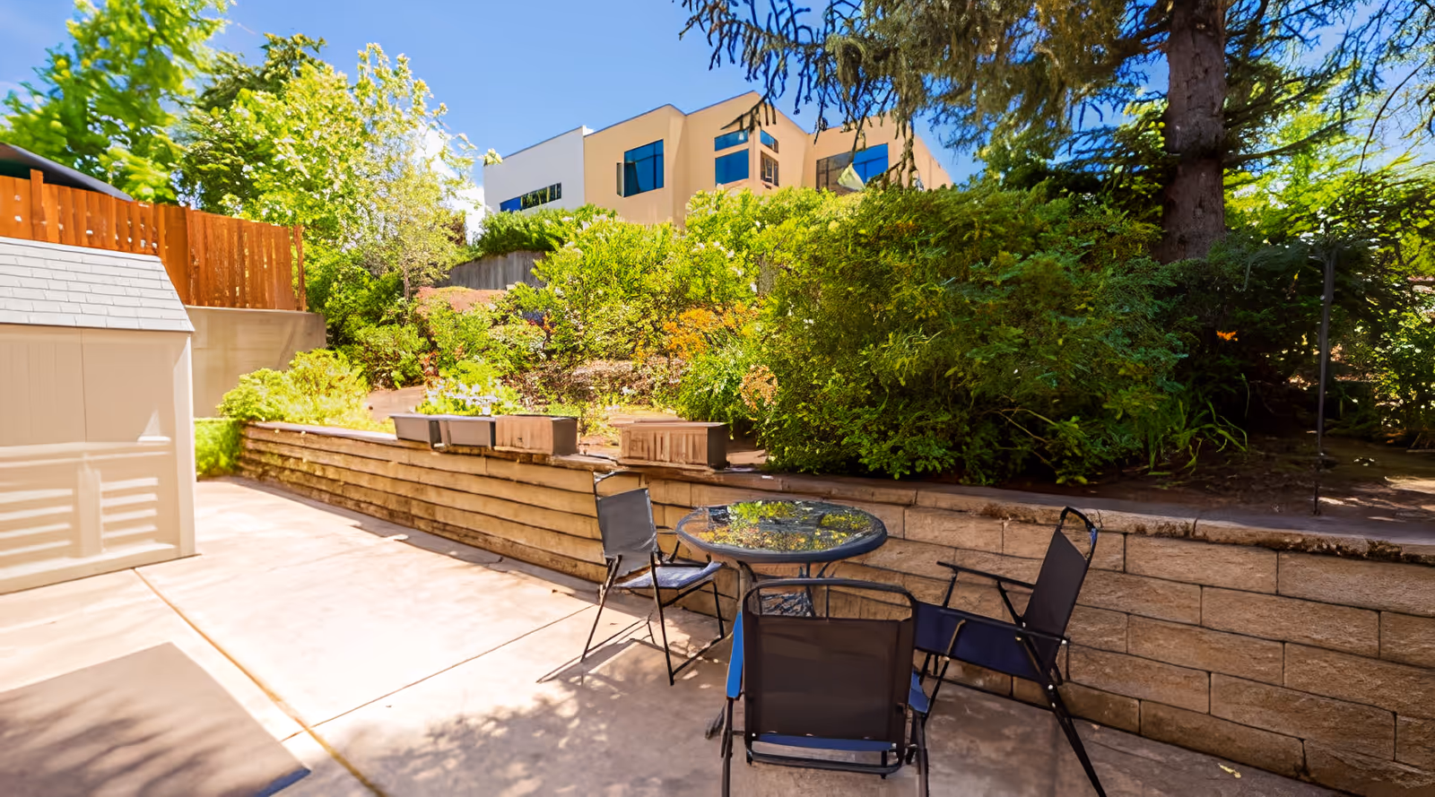 Outdoor patio area with a round glass table and three black chairs on a concrete surface, surrounded by a low stone retaining wall and lush green bushes and trees under a clear blue sky.