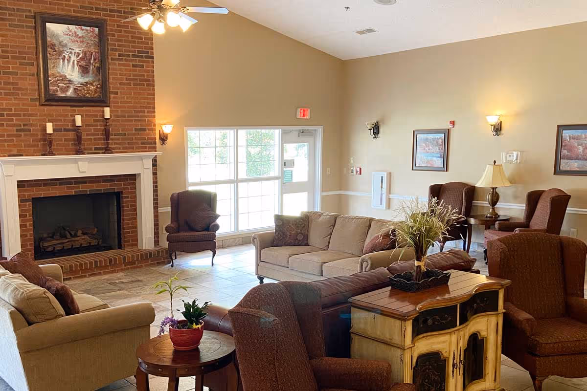 A cozy senior living facility common area with beige walls and tiled floor. The room features a brick fireplace with a white mantle, decorated with candles and a framed painting above. There are multiple upholstered armchairs and sofas arranged around the fireplace, with side tables holding plants and a lamp. Large windows and a glass door allow natural light to brighten the space.