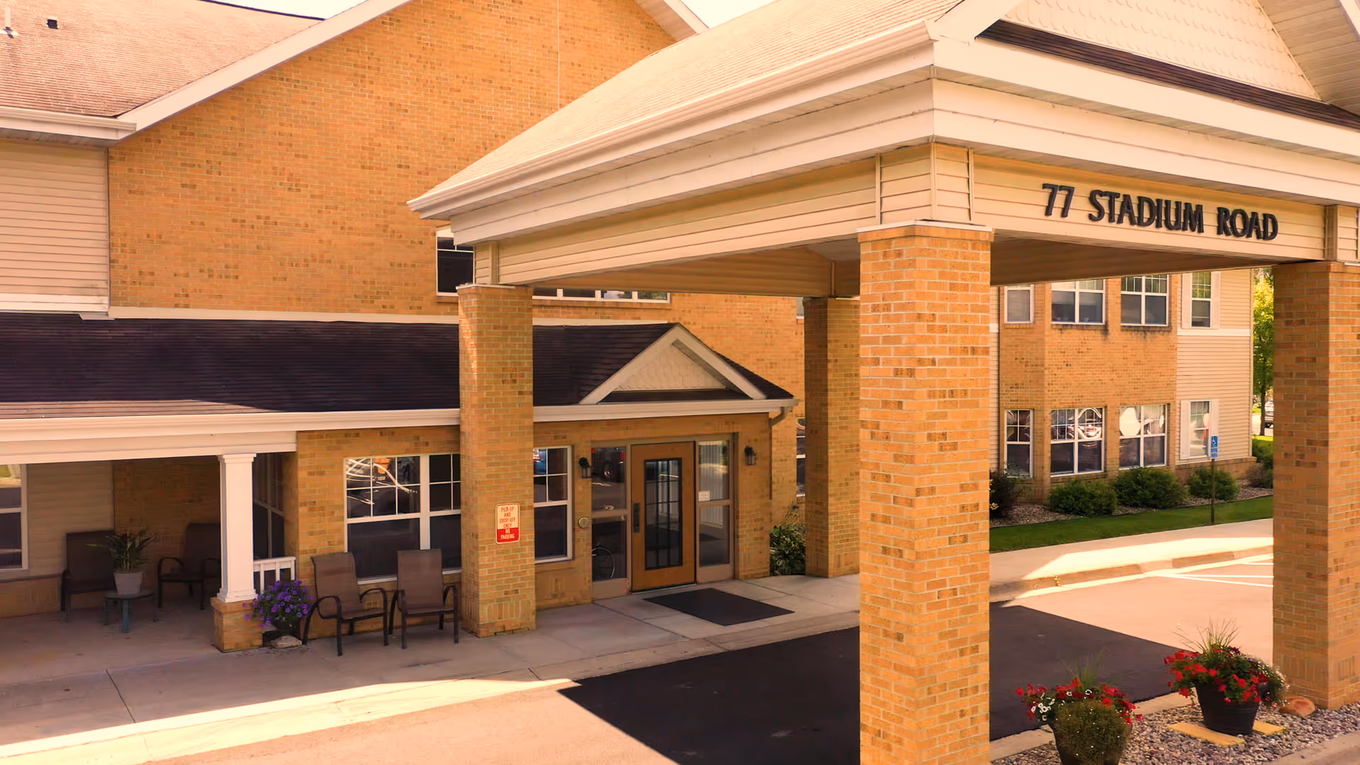 Entrance of a brick building with a covered driveway supported by brick pillars. The address '77 Stadium Road' is displayed on the front of the covered entrance. There are chairs and potted plants near the entrance door, and windows are visible on the building.